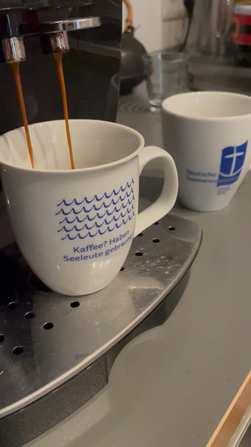 Unter einer Kaffeemaschine steht ein Becher. Auf dem steht Kaffee? Haben Seeleute gebracht. Dahinter steht ein Becher mit dem Logo der deutschen Seemannsmission.