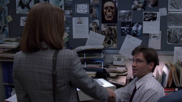 Fox Mulder's desk at the FBI. Weird paranormal things are everywhere. He sits there with glasses as he's meeting Dr. Dana Scully for the first time, her back is to us and we can just see her red hair and plaid checkered suit jacket.