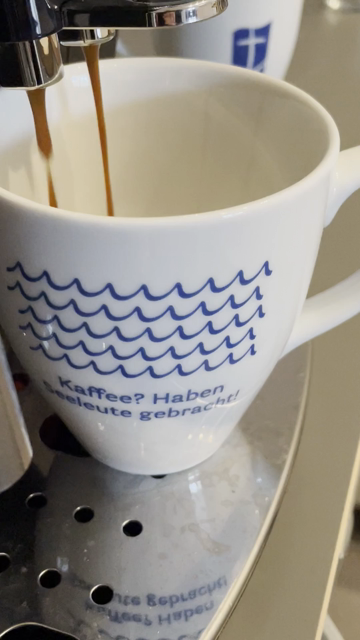 Auf einem weißen Kaffeebecher sind blaue bellen gemalt und es steht dort: Kaffee? Haben SeeLeute gebracht?
In den Becher läuft Kaffee aus einer Kaffeemaschine