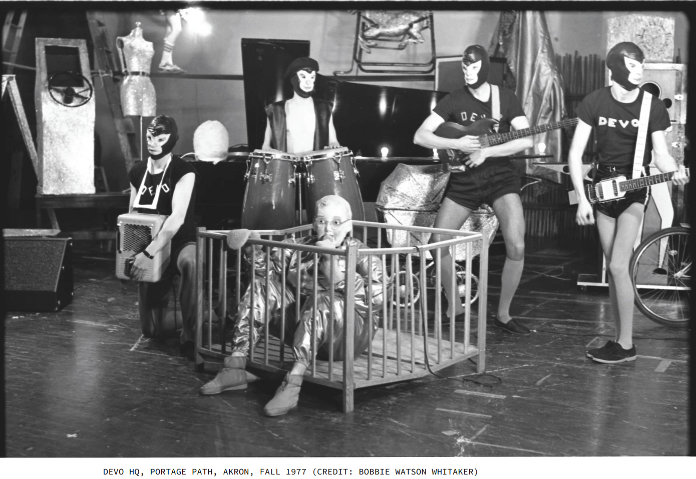 Black and white photo taken at DEVO's headquarters in Akron, Ohio. 4 band members wear rubber ape face masks, black t-shirts with DEVO in block letters, and black short shorts. In the foreground, one member wearing a metallic jumpsuit and a rubber "Booji Boy" mask sits in a baby crib. Text under photo: DEVO HQ, Portage Path, Akron, Fall 1977. Credit Bobbie Watson Whitaker. 