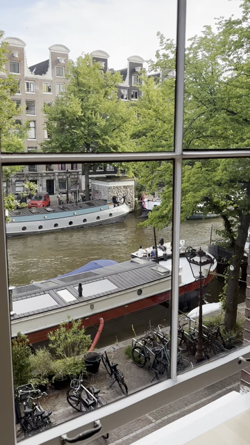 Durch das geteilte Fenster geblickt runter auf die Straße, an, der gracht, wo Fahrräder stehen, auf der Gracht liegen Hausboote und es fahren kleine Boote vorbei. Auf der anderen Seite stehen alte Grachten Häuser mit ihren Giebeln.