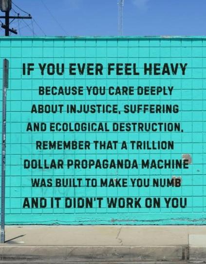 A bright turquoise wall features bold black text with a motivational message addressing feelings of heaviness due to caring about social and ecological issues. 

The text emphasizes the impact of a trillion-dollar propaganda machine, concluding with encouragement that it didn't succeed in overwhelming the reader.